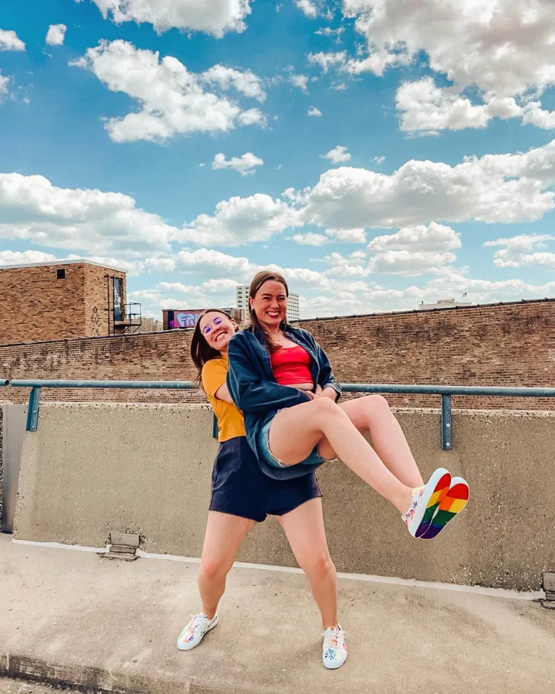 Two women pick one another up with a blue cloudy sky in the background